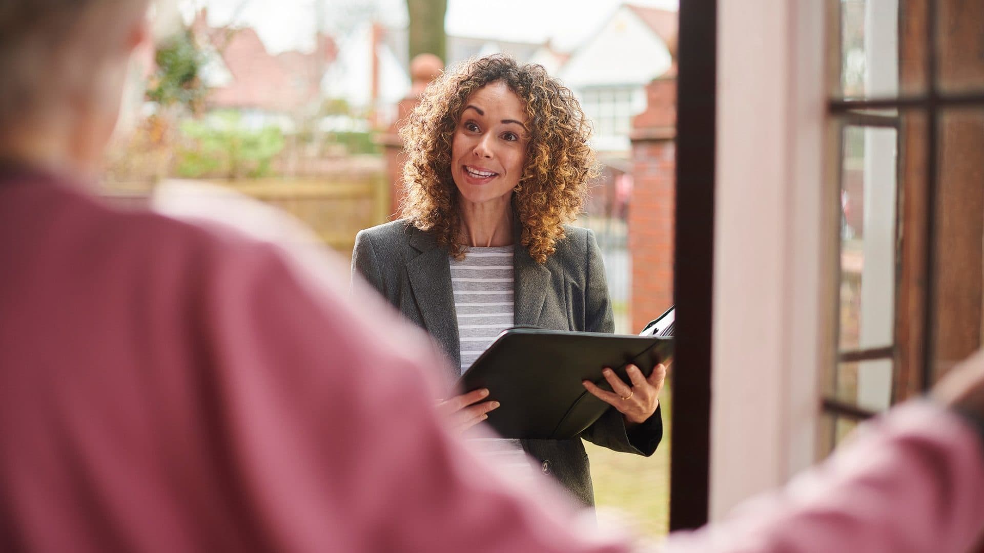 A woman holding a folder greets someone at the front door of a home
